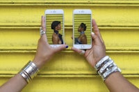 two hands holding up two iphones in front of a yellow wall